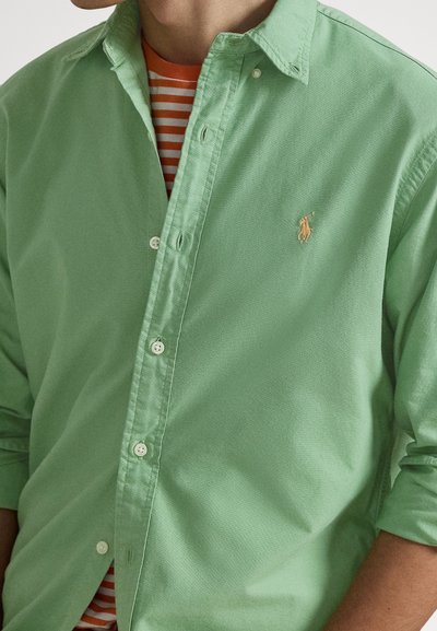 Ljusgrön skjorta i bomull med klassiskt krage. Bärs över en orange och vitrandig T-shirt; har en liten logga på bröstet.