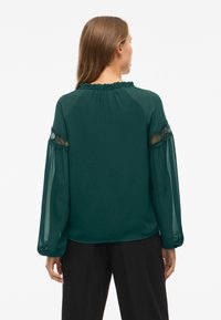Blusa verde in tessuto leggero, con maniche lunghe trasparenti e dettagli in pizzo, colletto arricciato e vestibilità morbida.