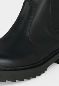 Gros plan d’une botte en cuir noire avec un panneau latéral élastique et une semelle en caoutchouc épaisse rainurée.