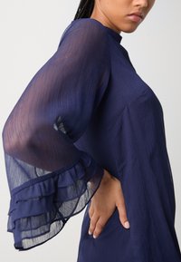 Navy blue dress with sheer, layered ruffle sleeves and a textured body. Features a high neckline and a fitted silhouette.