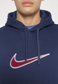 Man som bär en marinblå hoodie med en vinröd och vit broderad Nike swoosh-logotyp på bröstet mot en enfärgad bakgrund.