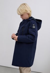 Blond kind in een marineblauwe hooded jacket met een logo-patch op de mouw, die de rits van de jas aanpast, staande tegen een effen muur.