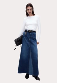 Denim-Maxirock mit einem Frontschlitz, kombiniert mit einem weißen Langarmshirt. Schwarzer Gürtel, übergroße schwarze Handtasche und schwarze Stiefeletten mit Absatz.