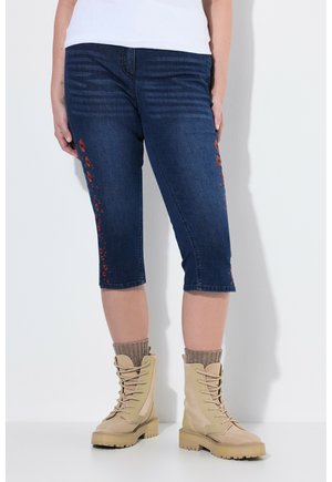 Person trägt dunkelblaue, verkürzte Jeans mit roter Blumenstickerei und beigen Schnürstiefeln mit braunen Socken vor weißem Hintergrund.
