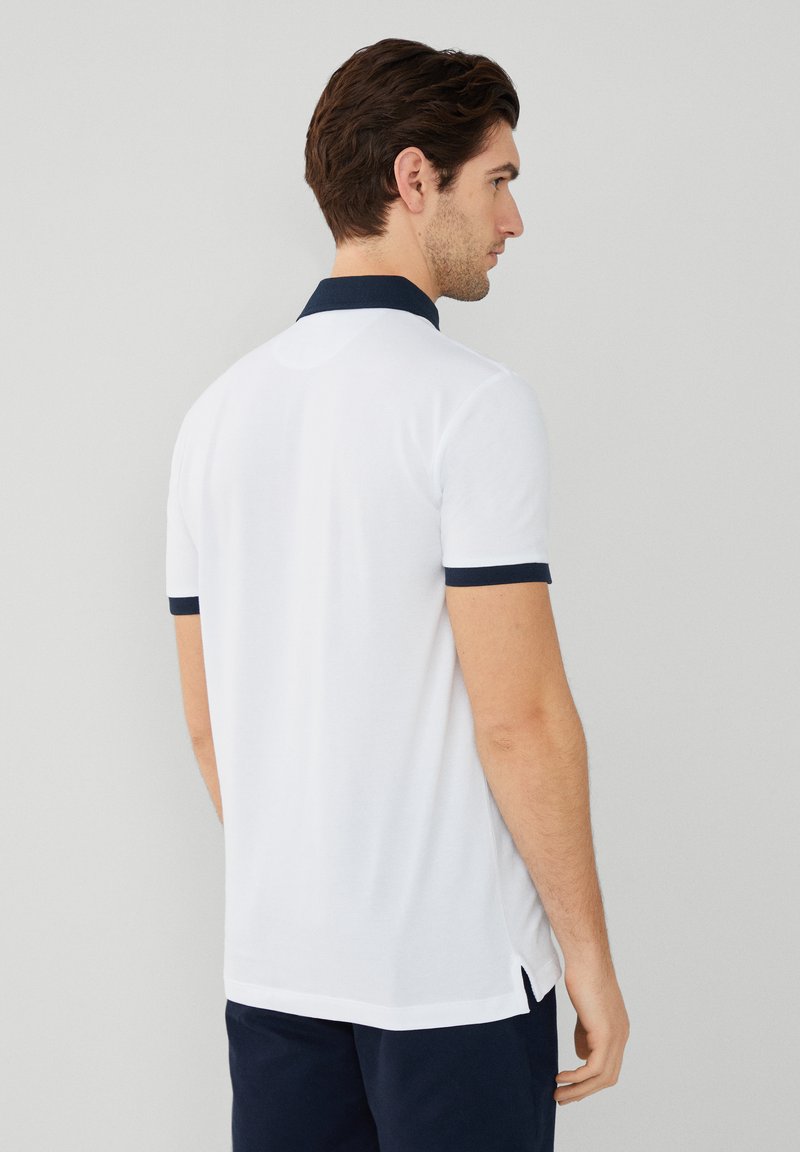 Polo shirt blanc avec des accents navy sur le col et les manches, manches courtes, fentes latérales, et une texture lisse. Modèle vu de dos.