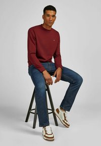 Bordeauxfarbenes Sweatshirt mit Rundhalsausschnitt, dunkle Jeans und weiße-braune Sneakers. Model sitzt auf einem schwarzen Metallhocker vor einem grauen Hintergrund.
