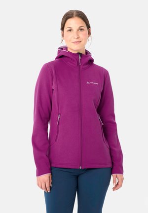 Donna che indossa una giacca viola con cappuccio e zip e pantaloni blu scuro, in piedi e rivolta in avanti su uno sfondo bianco semplice.