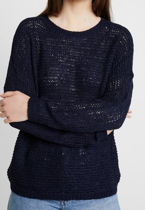 Person wearing a navy blue loosely knit sweater with a round neckline and light blue jeans, arms crossed over the chest.