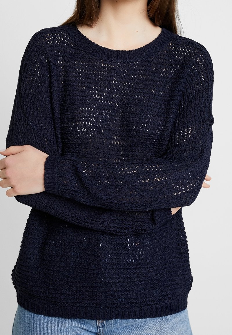 Personne portant un pull en maille lâche bleu marine à encolure ronde et un jean bleu clair, bras croisés sur la poitrine.