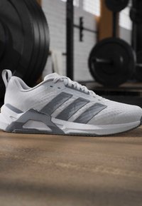 Baskets blanches et grises avec trois bandes, posées sur le sol d'une salle de sport avec des poids et du matériel flous en arrière-plan.