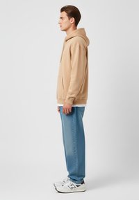 Felpa con cappuccio beige con tasca frontale, abbinata a jeans blu chiaro e sneakers bianche con accenti neri, vista di lato.
