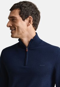 Marineblauwe sweater met ritssluiting, gemaakt van zacht materiaal, met een hoge kraag, minimalistisch ontwerp, klein geborduurd logo en leren ritstrekkertje.