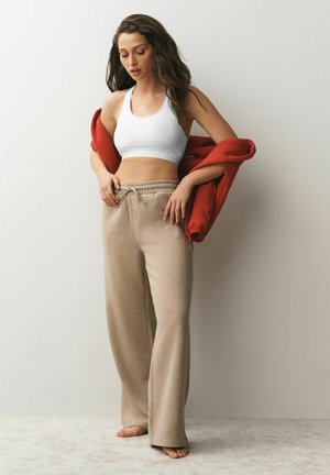 Frau in beigen Hosen mit weitem Bein und weißem Sport-BH hält eine rote Jacke und steht barfuß vor einer schlichten hellen Wand.