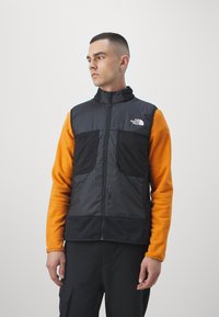 Black insulated vest with high collar, front zip, two large pockets, worn over an orange fleece top, against a plain white background.