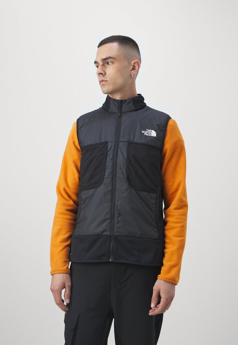 Black insulated vest with high collar, front zip, two large pockets, worn over an orange fleece top, against a plain white background.