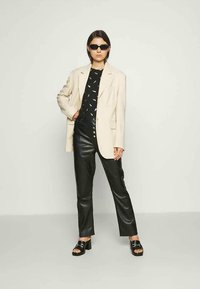 Mulher vestindo um blazer bege oversized, camisa preta estampada, calças de couro pretas de cintura alta, sandálias de salto pretas e óculos de sol pretos.