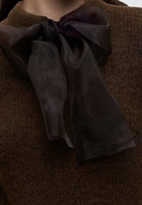 Nœud noir transparent noué autour du cou sur un pull en maille marron texturé.