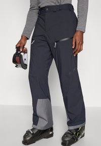 Παντελόνι για σκι σε σκούρο ναυτικό μπλε με γκρι λεπτομέρειες· διαθέτει τσέπες με φερμουάρ, φαρδιά ζώνη μέσης και ομαλή υφή. Φοριέται με μπότες για σκι.
