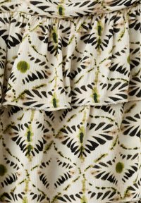 Tissu blanc avec des volants superposés présentant un motif floral abstrait vert et noir avec des dessins symétriques en forme de feuilles.