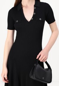 Robe noire côtelée avec un col en V, des manches courtes et cinq boutons décoratifs. Un sac à main noir à la forme arrondie avec un logo à la main.