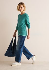 Turkos stickad tröja med rund halsringning, kombinerad med lösa blå denimjeans. Modellen håller en mörk denimväska och har på sig vita sneakers.