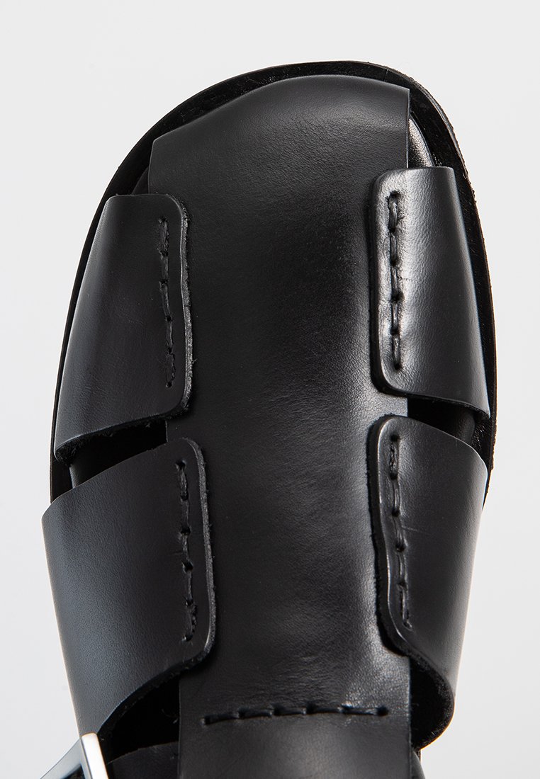 Zwarte leren sandaal met een platte zool, voorzien van twee brede bandjes en opvallende stiksels. Minimalistisch ontwerp met metallic hardware accenten.