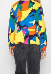 Färgglad blus med stora abstrakta mönster i blått, gult, grönt och rött, med långa ärmar och midjebälte. Bärs tillsammans med mörka jeans.