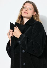Mladá žena s vlnitými blond vlasy, která má na sobě náušnice na kruhu a černý texturovaný kabát, drží límec oběma rukama.