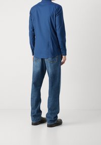 Niebieska koszula z długim rękawem z kołnierzykiem, zestawiona z luźnymi niebieskimi dżinsami i czarnym obuwiem; widoczny tylny szew i detale na dżinsach.