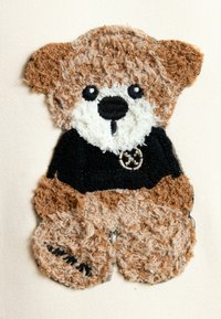Diseño de oso de felpa texturizado con pelaje marrón y crema. Lleva una camiseta negra con un emblema circular. Apariencia suave y esponjosa en todo su cuerpo.