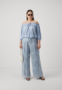 Top albastru deschis, cu umerii descoperiți, cu guler răsfirat, asociat cu pantaloni largi, cu model; modelul ține o geantă mică albă și poartă ochelari de soare.