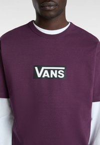 Fialové tričko s krátkým rukávem a černým logem "VANS" na hrudi, s kulatým výstřihem a bílou košilí s dlouhým rukávem pod ním.