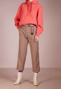 Koraalkleurige hoodie met trekkoord, geruite broek in oranje, bruin en zwart, gecombineerd met witte enkellaarsjes en een riem met de tekst "HUNDRED."