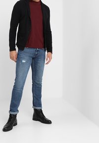 Musta napillinen neuletakki viininpunaisen paidan päällä, yhdistetty vaaleansinisiin repaleisiin farkkuihin ja mustiin nyörikenkiin. Yksinkertainen malli, rento istuvuus.