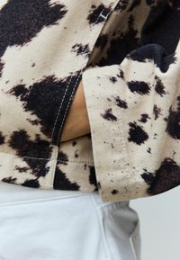 Beige tyg med svart stänkmönster, med en vikbar manschett, synlig sömnad och texturerad yta. Ställt mot vita byxor.