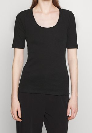 Kobieta nosząca dopasowany czarny top z krótkim rękawem i dekoltem w serek, stojąca na tle jasnego, jednolitego tła.