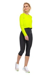 Neon gelbes langärmliges Croptop, kombiniert mit schwarzen Capri-Leggings und weißen Turnschuhen, mit bunten Akzenten und einem figurbetonten Design.