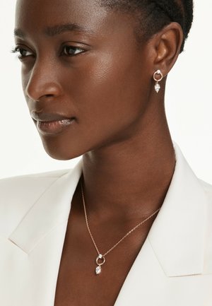 Femme portant des boucles d'oreilles et un collier assortis en or avec des pierres précieuses claires en forme de goutte, vêtue d'un blazer blanc.