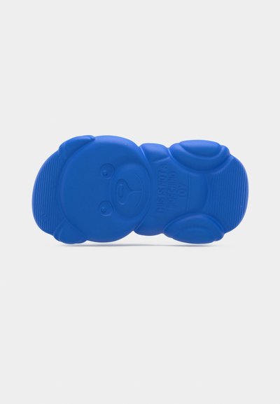 Μπλε λαστιχένια σόλα σε σχήμα αρκουδάκι με ανάγλυφες χαρακτηριστικές όψεις προσώπου και κείμενο που γράφει "ΑΥΤΟ ΔΕΝ ΕΙΝΑΙ ΠΑΙΧΝΙΔΙ MOSCHINO."