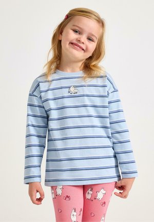 Ung pige smiler, iført en lyseblå langærmet skjorte med striber og en lille hundeapplikation samt pink leggings med hvide tegneserieflodheste og hjerter.