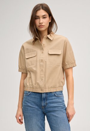 Frau trägt beige kurzärmliges Hemd mit Knopfleiste, elastischem Saum und zwei Brusttaschen, kombiniert mit blauen Jeans mit hoher Taille.