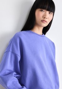 Sudadera púrpura con un corte relajado, cuello redondo y textura suave. La tela se ve lisa con detalles de costura visibles.