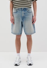 Pantalones cortos de mezclilla azul claro con un ajuste relajado, dobladillo deshilachado y cinco bolsillos. Llevados con zapatillas deportivas blancas y calcetines blancos visibles.