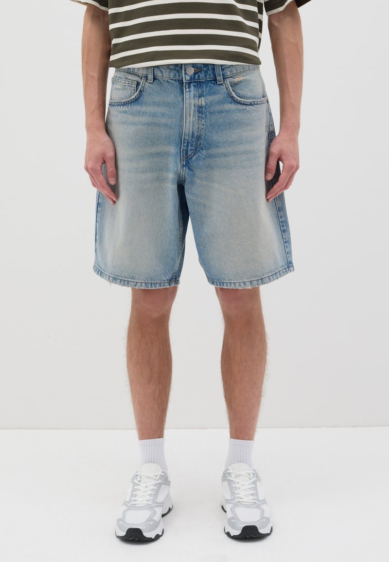 Pantalones cortos de mezclilla azul claro con un ajuste relajado, dobladillo deshilachado y cinco bolsillos. Llevados con zapatillas deportivas blancas y calcetines blancos visibles.