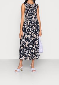 Eine Frau steht in einem blumigen marineblauen Kleid und hält eine weiße Tasche. Sie trägt hellviolette Absatzsandalen vor einem schlichten Hintergrund.