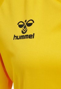 Maglia sportiva gialla realizzata in tessuto testurizzato, con un logo "hummel" nero e un design di ape sul lato superiore sinistro.