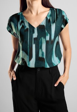 Vrouw die een V-hals blouse met een abstract patroon in teal, zwart en lichtblauw draagt, met korte mouwen, en deze in een zwarte broek gestopt heeft, handen in de zakken.