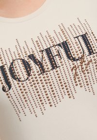 T-shirt beige clair à manches courtes arborant un graphisme "JOYFUL" en bleu foncé et des clous décoratifs en rose doré disposés en motif linéaire.