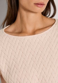 Der Nacken und das untere Gesicht einer Frau, die einen hellrosa strukturierten Strickpullover mit einem Bootsausschnitt trägt.