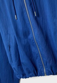 Modrá lehká bunda se stříbrným zipem, modrými šňůrkami s kovovými konci a elastickým pasem, zobrazená zblízka.